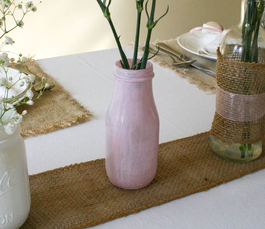 Pink vase sitting on burlap ribbon with flowers stalks visible.
