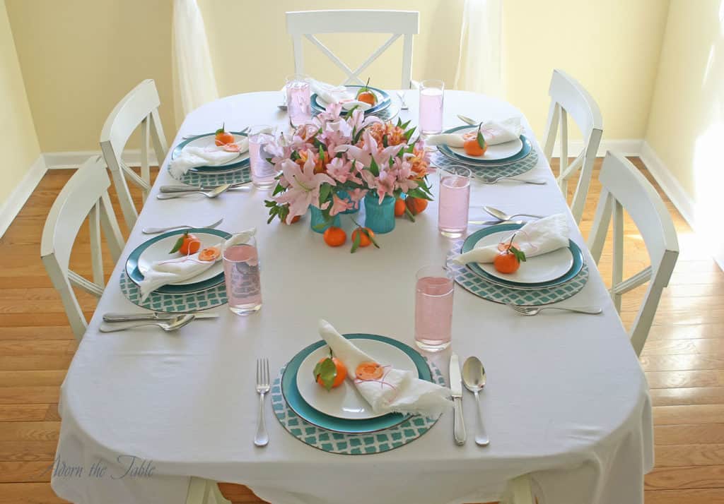 Mother's Day table setting with teal vases in the middle, and pink and orange flowers. Teal and white chargers, teal plates, white napkins with DIY orange napkin rings.