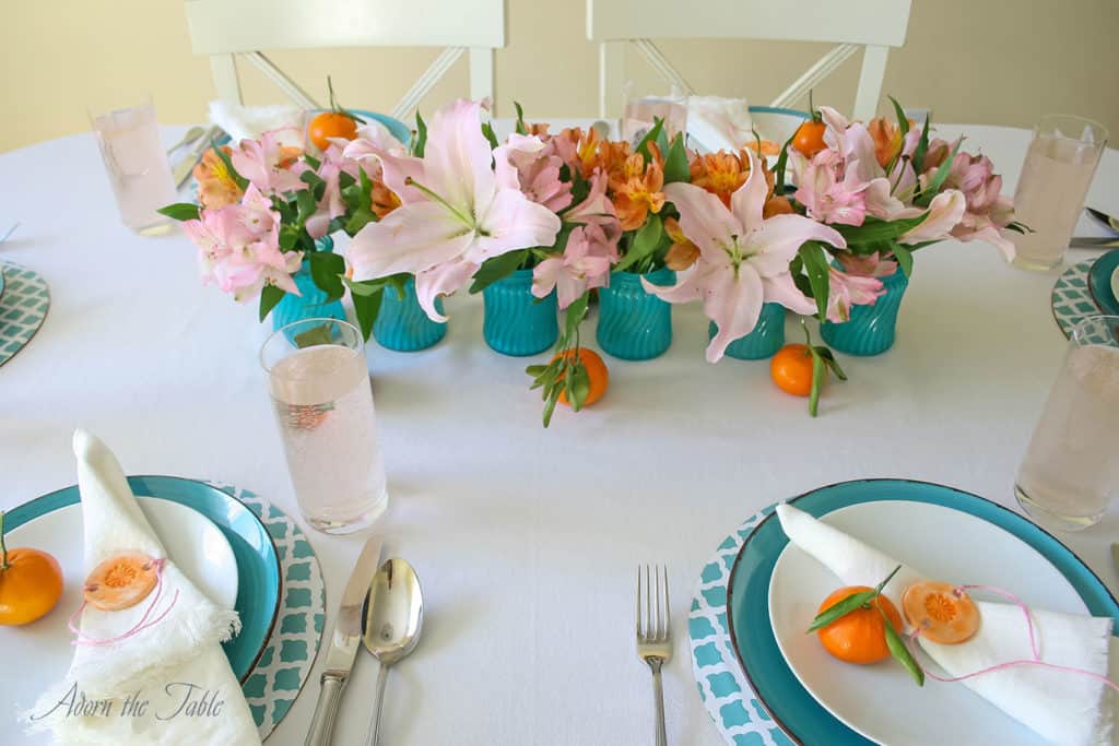 Centerpieces three ways - table setting straight on view of six teal vases with pink and orange flowers lined up on the middle of the table. 