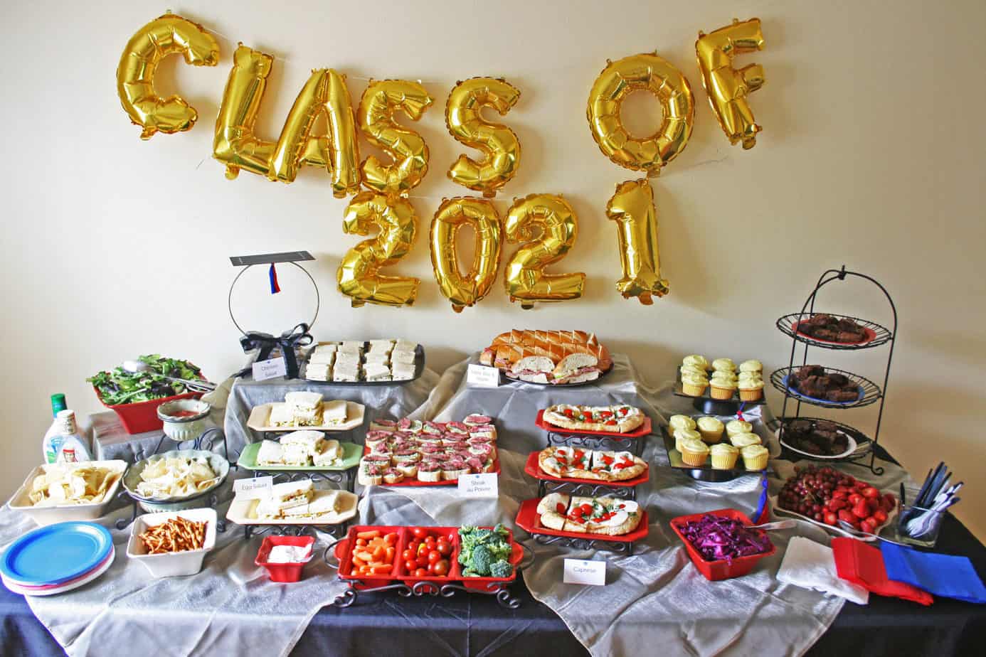 Graduation buffet table with elevated food, a graduation cap centerpiece and Class of 2021 sign.
