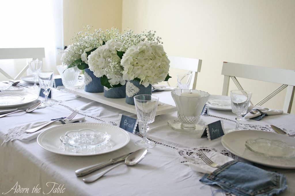 Side view of table. With denim covered vases on a white wooden serving tray, on a white tablecloth. Denim place cards and napkin holders made from old jeans. 