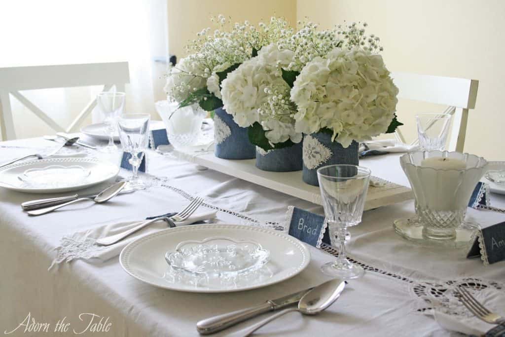 Side view of table. With denim covered vases on a white wooden serving tray, on a white tablecloth.