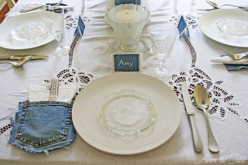 Place setting with white lace tablecloth, white and clear plates and denim place card.