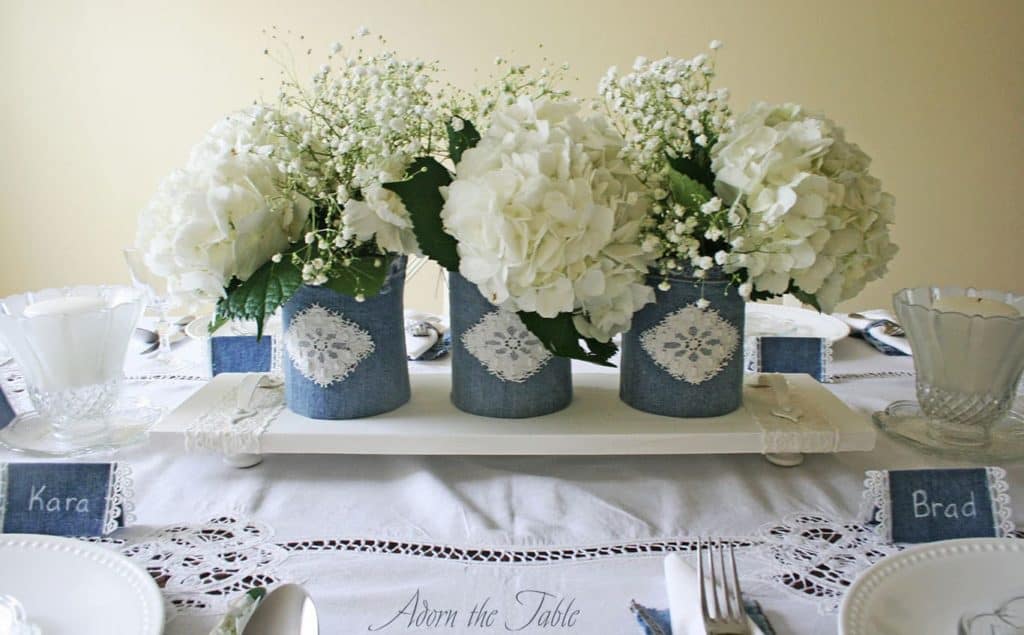 Side view of denim and white lace centerpiece. Denim vases sitting on white wooden tray with white flowers, on a white tablecloth.