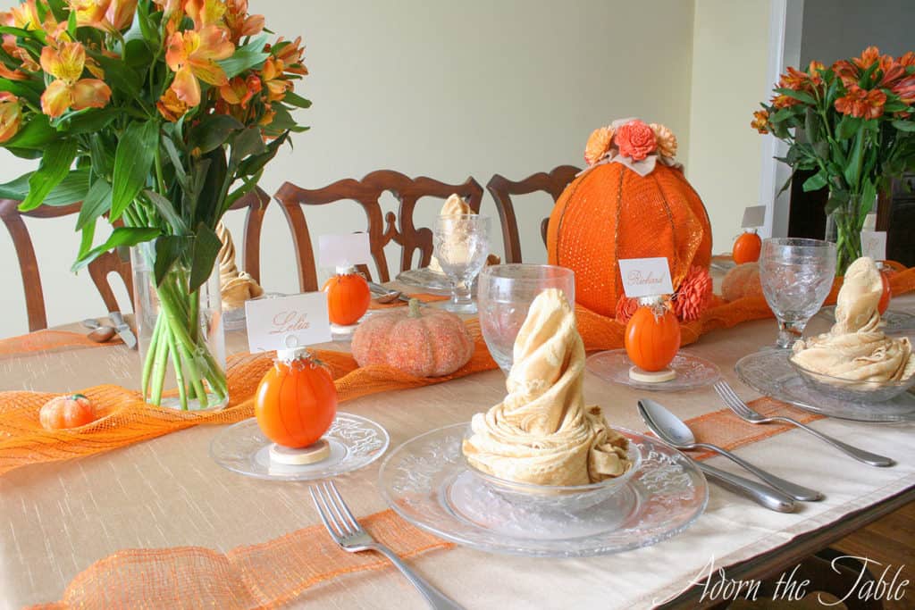 Pumpkin table setting view from end of the table. Clear plates with champagne colored napkins, diy pumpkin place card holders, a large pumpkin centerpiece and orange flowers.