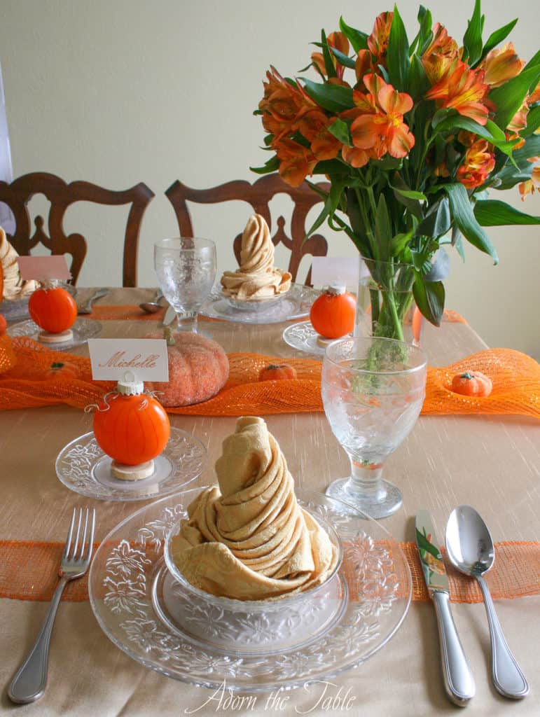 Table setting for Autumn with orange flowers, orange pumpkin placecard holders and champagne napkins on clear plates.