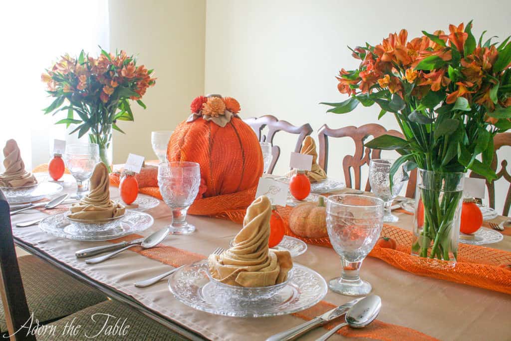 Pumpkin table setting view from end of the table. Clear plates with champagne colored napkins, diy pumpkin place card holders, a large pumpkin centerpiece and orange flowers.