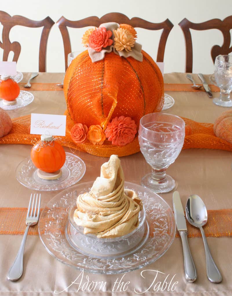 Table setting for Fall with orange pumpkin centerpiece, orange pumpkin place card holders and champagne napkins on clear plates.