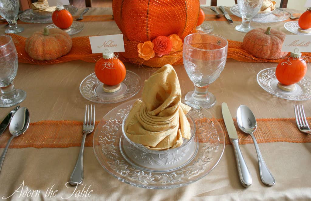 Place setting for autumn table with gold napkin on clear plates with a pumpkin place card holder. 