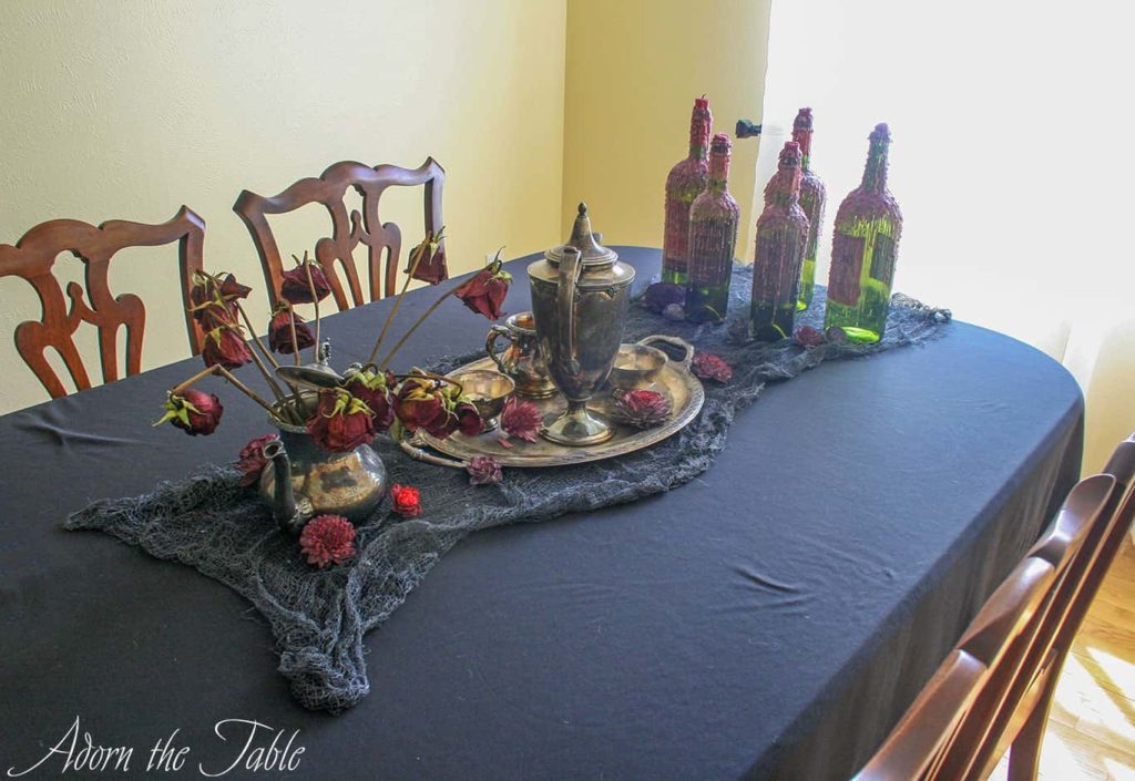 Black tablecloth with tarnished silver teapot set, dead flowers and candle wax covered wine bottles.