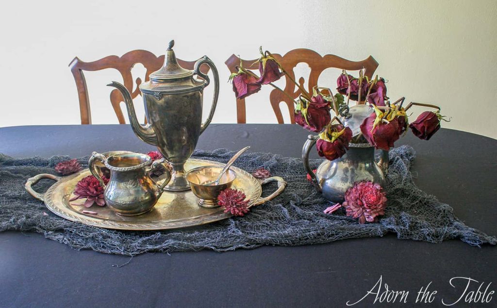 Black tablecloth with tarnished silver teapot set and dead flowers