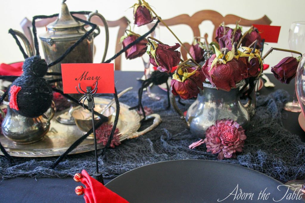 Spider place card holder with red name card. Silver teapot filled with dead roses.