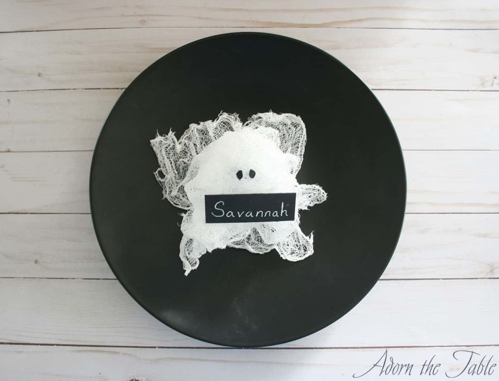 Overhead view of Halloween DIY Cheesecloth Ghost Place Card Holder with black eyes on a black plate holding a black name card