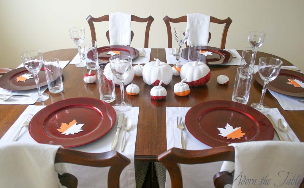 Simple modern Thanksgiving day tablescape showing white placemats, red chargers and white pumpkins.