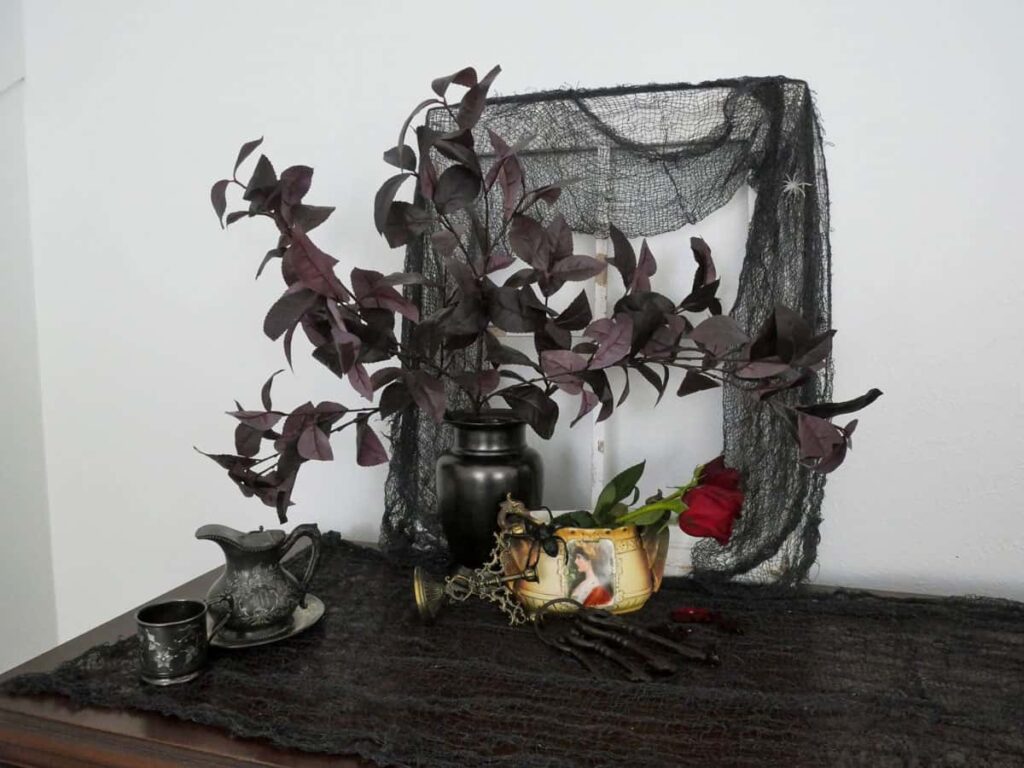 A wooden picture frame draped with grey creepy cloth and a black vase with deep plum stems in front of it.