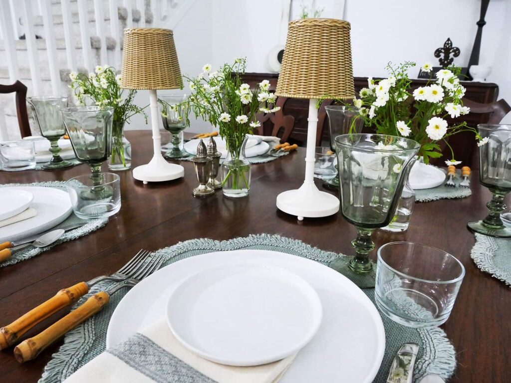 Tablescape with white plates, light grey/green placemat and portable lamps with wicker shades.